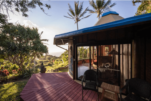 A cozy cabin with a blue roof, surrounded by lush greenery and palm trees, overlooking a scenic landscape at sunset.