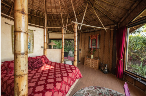 Cozy bamboo hut interior with a red patterned bedspread, wooden accents, and large windows overlooking greenery.