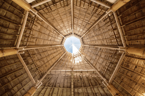 View from below of a circular, thatched roof with a central skylight, showcasing wooden beams and natural materials.