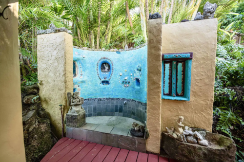 A colorful outdoor shower area with blue walls, decorative stones, and tropical plants surrounding it.