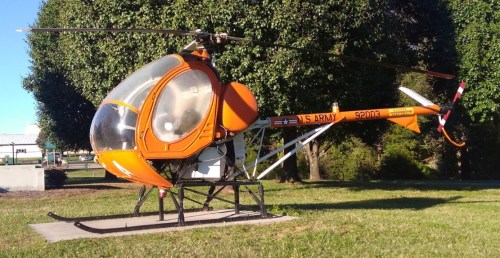 An orange military helicopter on display in a grassy area, with trees and a building in the background.