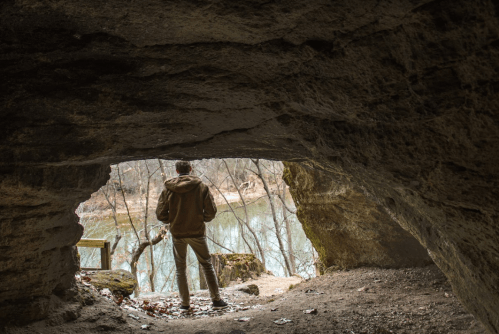 A person stands in a cave entrance, looking out at a serene river surrounded by trees.