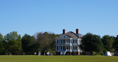 A historic white plantation house surrounded by green grass and trees, with people gathered outside on a clear day.