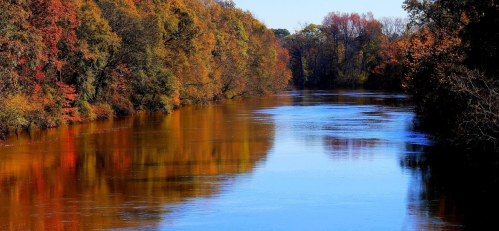 A serene river reflects vibrant autumn foliage along its banks, showcasing rich colors of orange, red, and yellow.