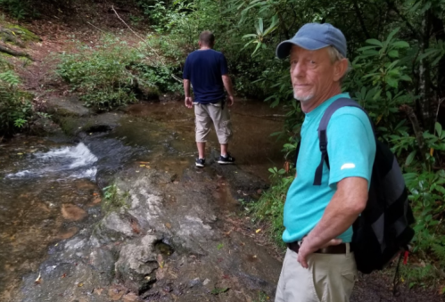 Two men near a stream in a wooded area; one stands in the water while the other poses for the photo.