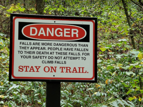 Sign warning about the dangers of falls, advising to stay on the trail for safety.