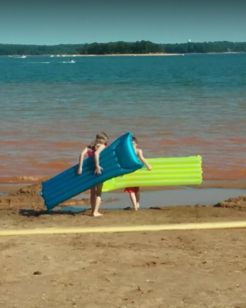 Two children carry colorful inflatable rafts along a sandy beach by a lake on a sunny day.