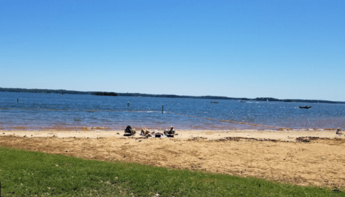 A sandy beach by a lake with people relaxing and boats in the water under a clear blue sky.