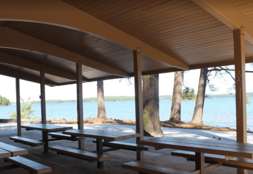 A covered picnic area with wooden tables overlooking a serene lake and trees in the background.