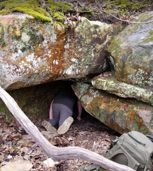 A person crawls into a rocky cave, surrounded by moss and fallen leaves, with a backpack nearby.