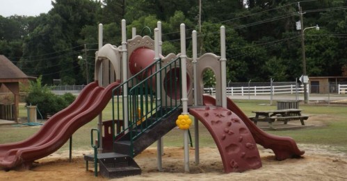 Colorful playground structure with slides, climbing features, and a sandbox, set in a grassy area with trees in the background.