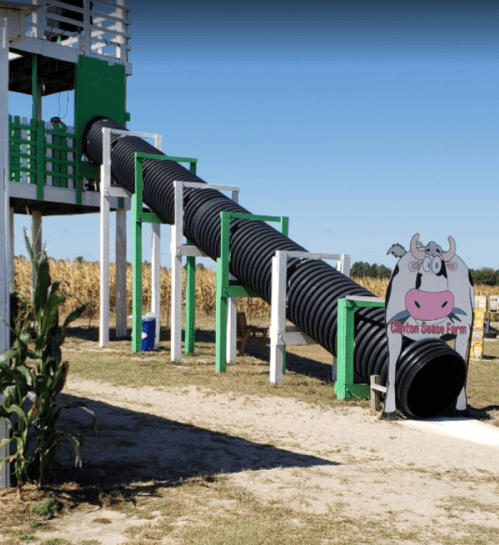 A long black slide extends from a green structure at Clinton Sausage Farm, with a cartoon cow sign nearby.