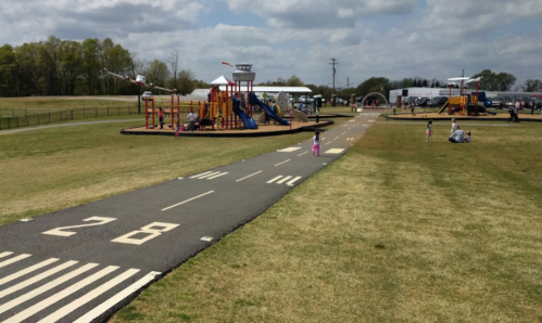 A playground with colorful equipment and a road-like path, children playing on the grass under a cloudy sky.