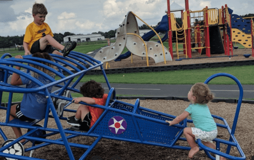 Children playing on a blue climbing structure at a playground, with slides and other equipment in the background.