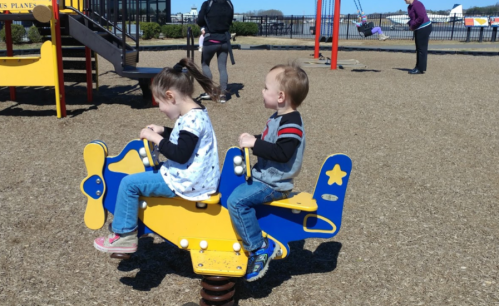 Two children play on a yellow airplane spring rider at a playground, with adults in the background.