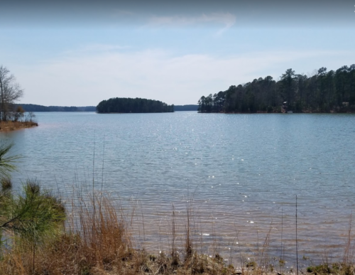 A serene lake scene with calm water, distant islands, and trees lining the shore under a clear blue sky.