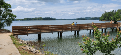 A wooden pier extends over calm water, with people enjoying the view and greenery surrounding the scene.