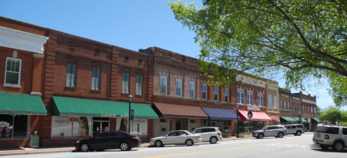 A row of historic brick buildings with green awnings and parked cars along a sunny street.