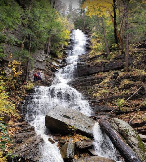 A cascading waterfall flows down rocky terrain, surrounded by trees with autumn foliage. Two hikers stand nearby.