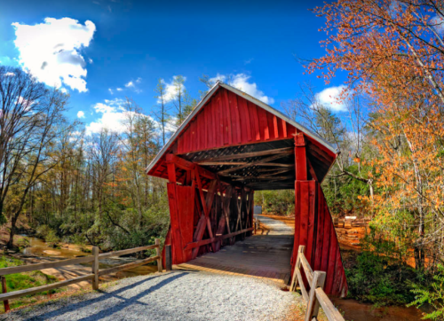 A red covered bridge spans a small creek, surrounded by trees and a clear blue sky with scattered clouds.