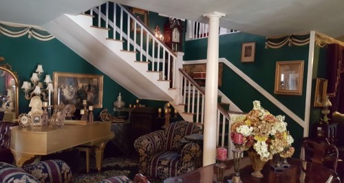 A vintage living room with green walls, a grand piano, floral sofas, and a staircase adorned with decorative items.