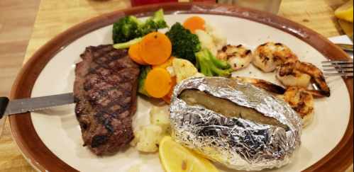 A plate featuring grilled steak, shrimp, mixed vegetables, and a baked potato wrapped in foil.
