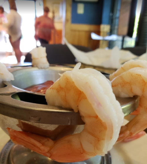 A close-up of shrimp in a metal bowl, with a blurred background of people in a restaurant.
