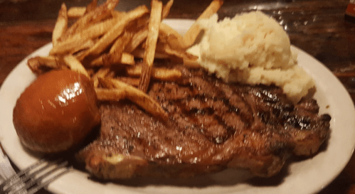 A plate featuring a grilled steak, crispy fries, creamy mashed potatoes, and a dinner roll.
