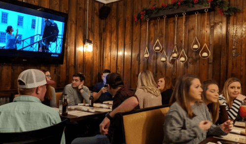 A cozy restaurant scene with diners at tables, a TV showing a movie, and wooden walls decorated for the holidays.