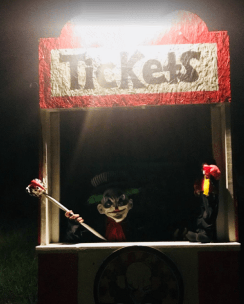 A creepy clown in a ticket booth at night, holding a scepter and surrounded by dark shadows.