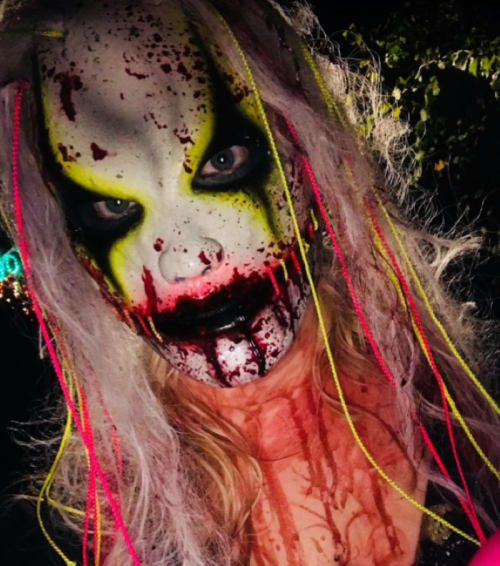 A person in a creepy mask with bright colors, long hair, and fake blood, set against a dark background.