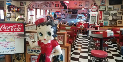 A retro diner interior featuring a Betty Boop statue, checkered floors, vintage decor, and classic cars in the background.