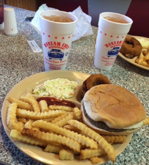 A plate with a burger, fries, coleslaw, and a hushpuppy, alongside two cups of soda on a table.