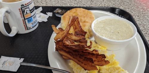 A breakfast plate with scrambled eggs, bacon, a biscuit, and a cup of grits, alongside a coffee mug and condiments.