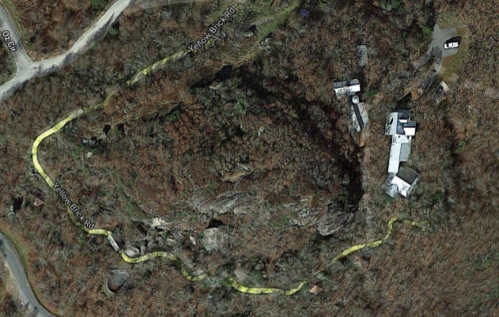 Aerial view of a winding road surrounded by wooded terrain, with several buildings visible along the path.