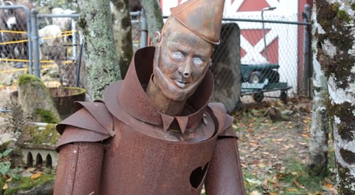 A rusty, humanoid sculpture with a conical hat, set in a garden with trees and a fence in the background.