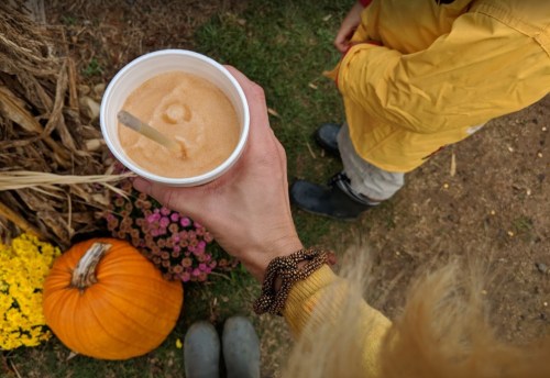 A hand holds a cup of orange drink above a pumpkin and flowers, with a child in a yellow jacket nearby.