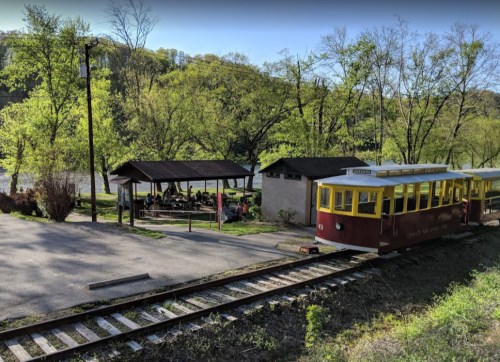 A vintage train on tracks near a picnic area surrounded by trees and a river in a scenic outdoor setting.