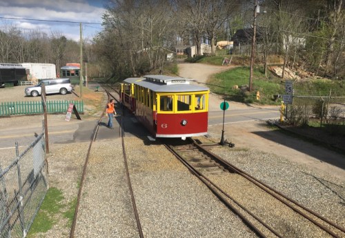 A red and yellow train car approaches a crossing, with a worker signaling and trees in the background.
