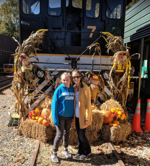 Two women pose in front of a decorated train with pumpkins, hay, and scarecrows, celebrating a fall theme.