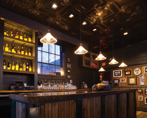 A cozy bar interior with wooden accents, shelves of liquor, and warm pendant lighting.