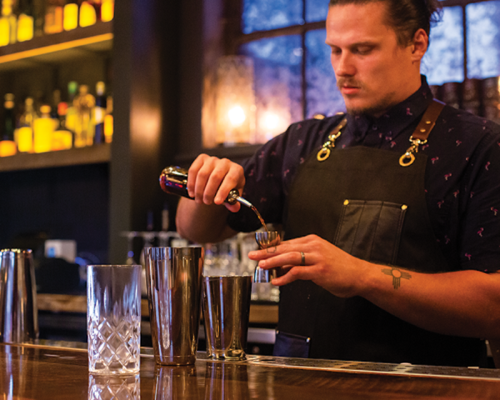 A bartender in an apron pours a drink into a jigger at a stylish bar with shelves of liquor in the background.