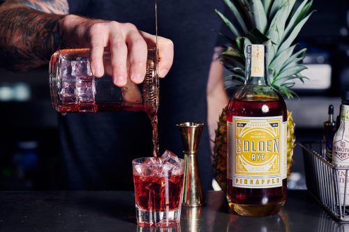 A bartender pours a red cocktail over ice into a glass, with a bottle of Golden Rye and a pineapple in the background.