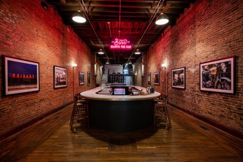 A rustic bar with a neon sign, surrounded by brick walls and framed photographs, creating a warm, inviting atmosphere.