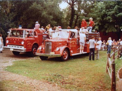 Two vintage fire trucks parked at an outdoor event, with people gathered around and climbing on one of the trucks.