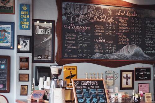 Chalkboard menu in a coffee shop featuring drinks, prices, and surf-themed decor on the walls.