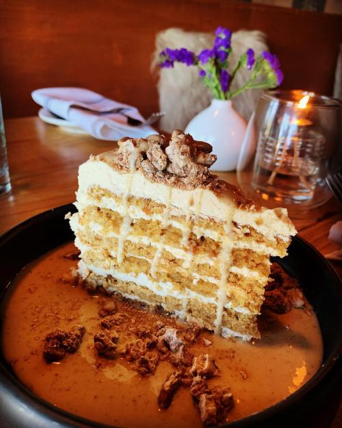A slice of layered cake topped with crushed nuts, served in a pool of sauce, with a cozy restaurant setting in the background.