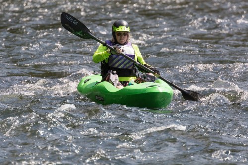 A person in a bright green kayak paddles through rippling water, wearing a helmet and sunglasses.