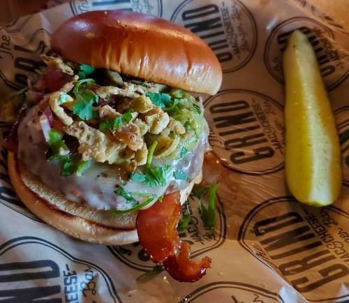A delicious burger with toppings, including bacon, onions, and herbs, served with a pickle on the side.