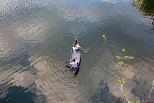 Aerial view of two people in a canoe on calm water, surrounded by reflections of clouds and trees.